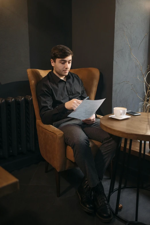 a man sitting in a chair reading a piece of paper, wearing black modern clothes, coffee shop, hasbulla magomedov, he is about 20 years old | short