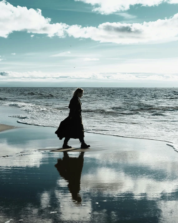 a woman walking on a beach next to the ocean, black reflect robe, non-binary, jamie campbell bower, of letting go