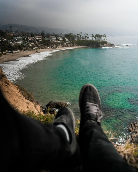 a person sitting on top of a cliff next to the ocean, sneaker photo, beach trees in the background, lgbtq, beach is between the two valleys