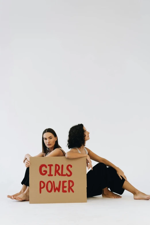 two women sitting next to each other holding a sign, by Olivia Peguero, trending on pexels, feminist art, hybrid of gal gadot, no power, girl standing, - 12p