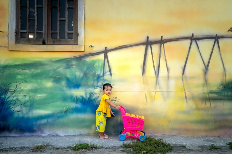 a little boy that is standing in front of a wall, inspired by Steve McCurry, pexels contest winner, street art, shopping cart, ao dai, pink and yellow, square