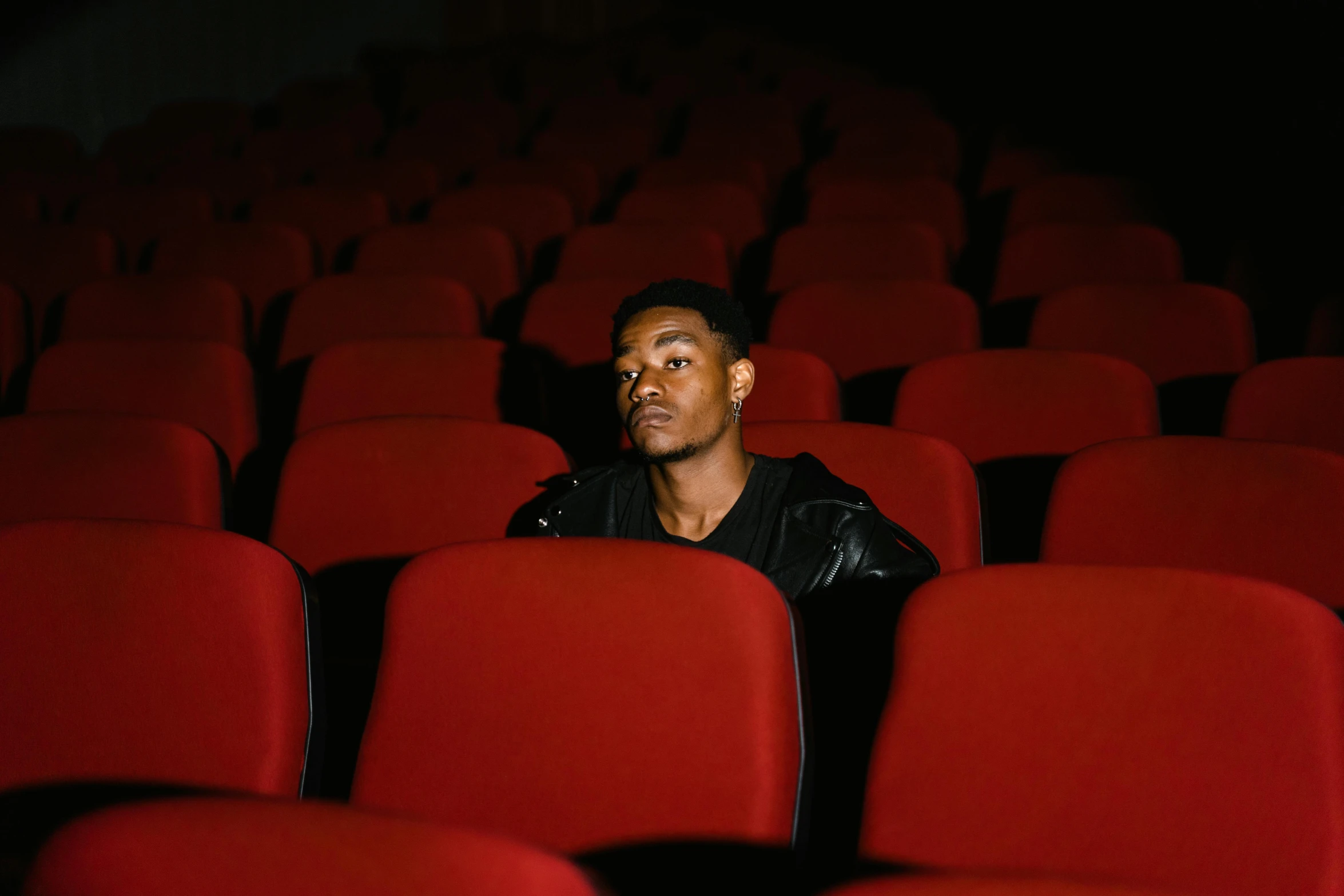 a man sitting in the middle of rows of red chairs, an album cover, pexels, realism, mkbhd, sitting in a movie theater, black teenage boy, ignant