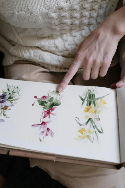 a woman holding a book with watercolor flowers on it, by Miyamoto, trending on pexels, herbarium page, finger painting, color image, patchy flowers