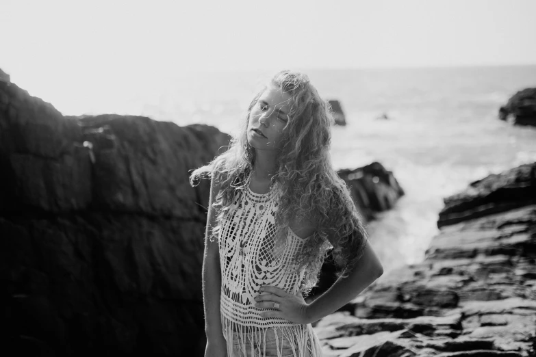 a woman standing on a rocky beach next to the ocean, a black and white photo, by Caroline Mytinger, pexels, fine art, long braided curly blonde hair, hippie and boho fashion 1970s, portrait shot 8 k, in clothes! intricate
