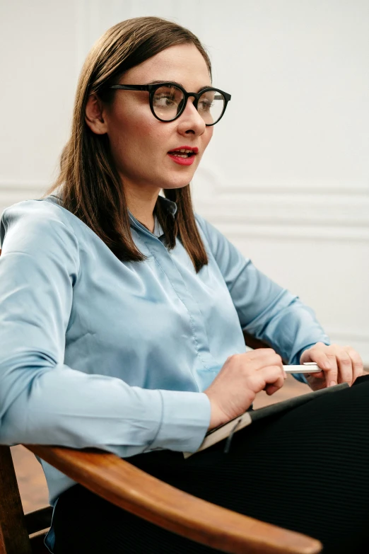 a woman sitting in a chair using a laptop computer, wearing square glasses, giving an interview, looking serious, 2019 trending photo