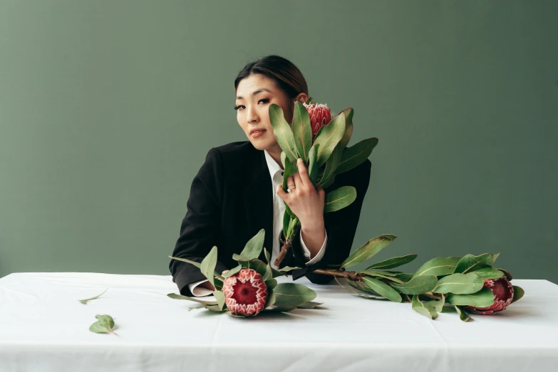 a woman sitting at a table with a bunch of flowers, an album cover, inspired by Ruth Jên, ikebana, wearing green suit, in front of white back drop, zeen chin and farel dalrymple