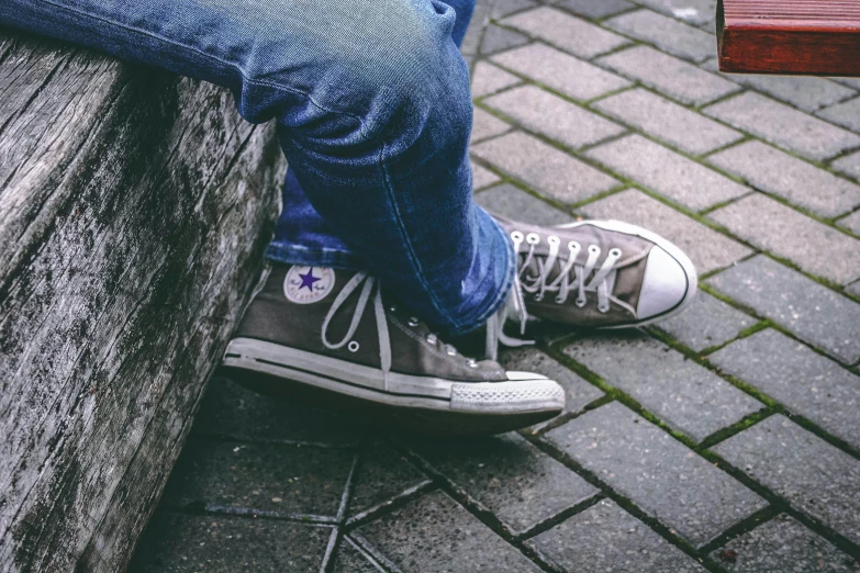 a person sitting on top of a wooden bench, a picture, blue jeans and grey sneakers, converse, gray skin. grunge, wikimedia commons