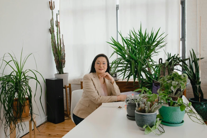 a woman sitting at a table surrounded by potted plants, inspired by helen huang, in office, louise zhang, plain background, looking towards camera