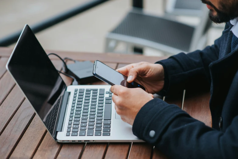 a man sitting at a table with a laptop and a cell phone, by Carey Morris, trending on pexels, outside, highly technical, typing on laptop, avatar image