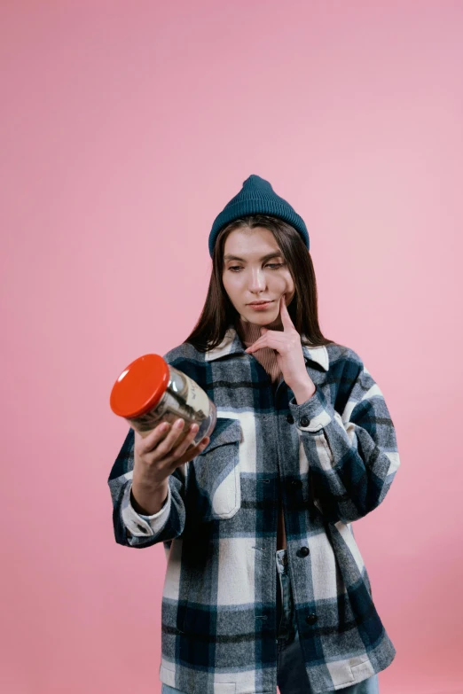 a woman standing in front of a pink background, by Julia Pishtar, trending on pexels, antipodeans, picking up a can beans, beanie, stoner, head in a jar