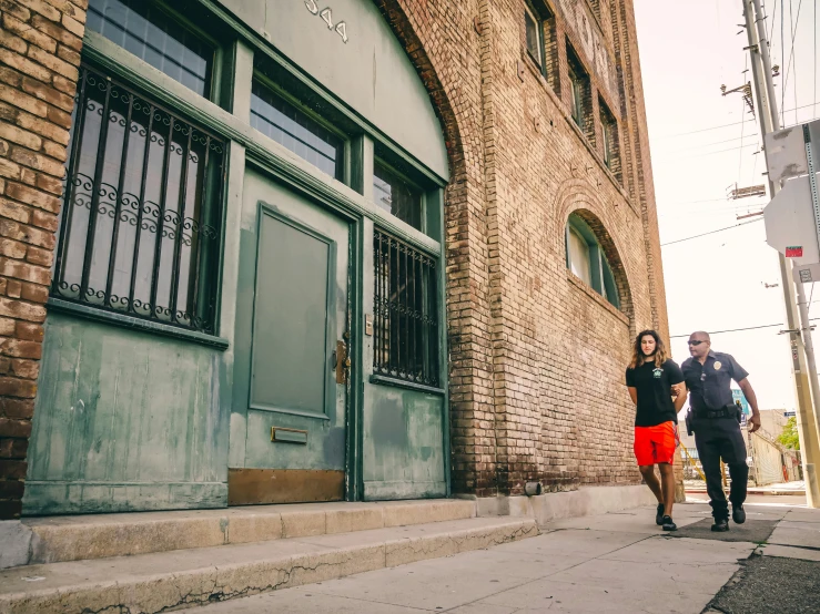 a man and a woman walking down a sidewalk, a photo, unsplash, art nouveau, brick building, los angeles ca, art deco factory, leaning on door