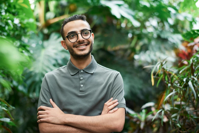 a man standing in a garden with his arms crossed, inspired by Ismail Gulgee, pexels contest winner, hurufiyya, jewish young man with glasses, in a dark green polo shirt, avatar image, middle eastern