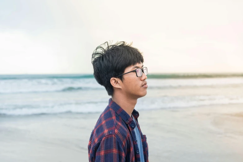 a man standing on top of a beach next to the ocean, a picture, inspired by Kim Eung-hwan, pexels, realism, in square-rimmed glasses, male teenager, profile image, thoughtful )