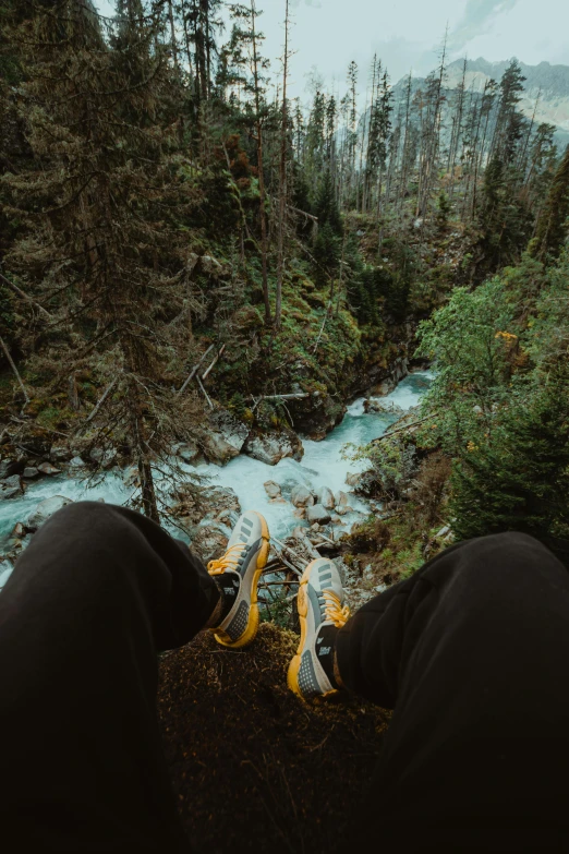 a person sitting on top of a mountain next to a river, legs taking your pov, hanging trees, sneaker photo, british columbia