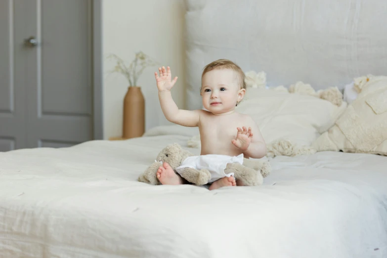 a baby sitting on a bed with a teddy bear, natural hands and arms, with arms up, linen, thumbnail