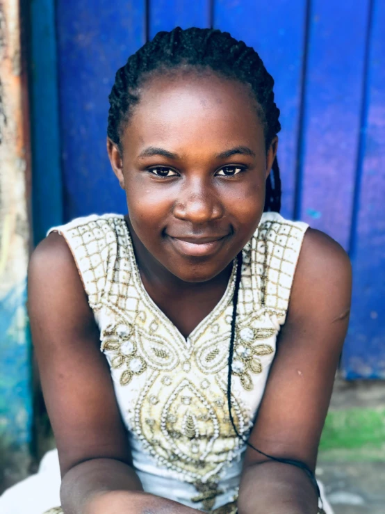 a young girl sitting in front of a blue door, dark skinned, pictured from the shoulders up, lovingly looking at camera, headshot profile picture