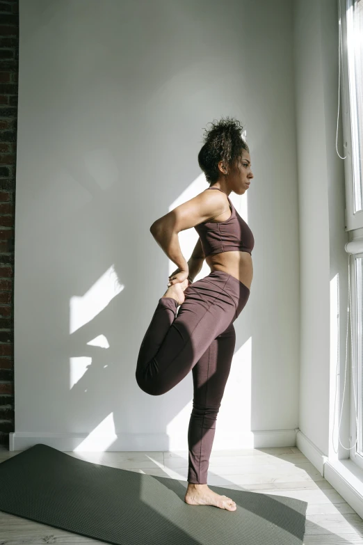 a woman standing on a yoga mat in front of a window, by Jessie Algie, pexels contest winner, arabesque, leggings, ashteroth, maroon, hero pose