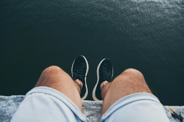 a man sitting on top of a rock next to a body of water, pexels contest winner, realism, gray shorts and black socks, focus on sneakers only, head straight down, vertical wallpaper