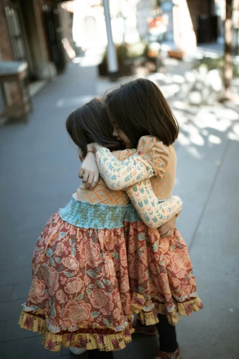 two little girls hugging each other on a sidewalk, unsplash, patterned, lightly dressed, handcrafted, trending photo