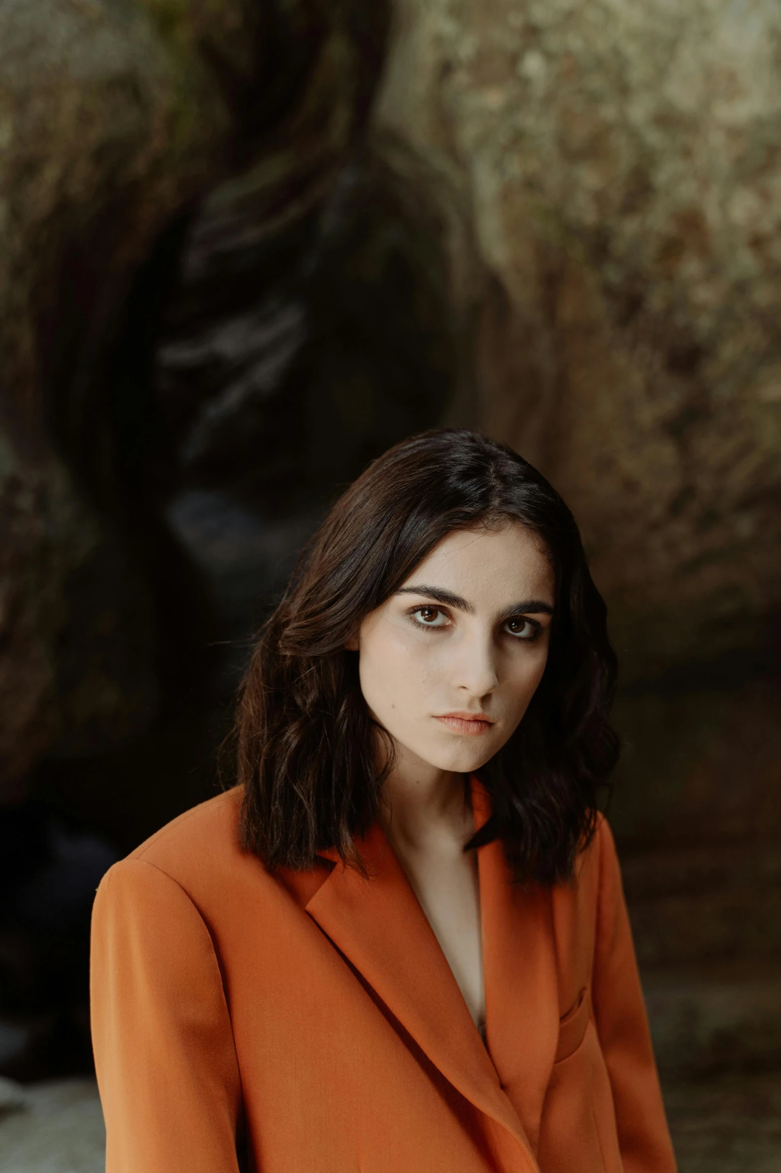 a woman standing in front of a large rock, an album cover, by irakli nadar, trending on pexels, photorealism, orange jacket, winona ryder, handsome girl, girl in suit