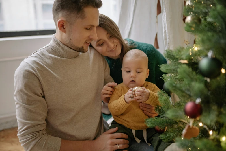 a man and woman holding a baby near a christmas tree, pexels, symbolism, profile image, brown, husband wife and son, gif