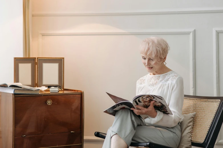 a woman sitting in a chair reading a book, a portrait, by Ruth Simpson, pexels contest winner, looking at the treasure box, white-haired, magazine photo, a very macular woman in white