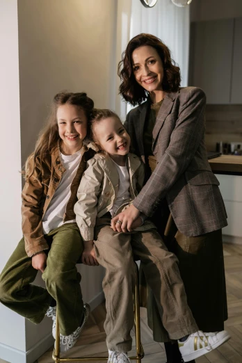 a woman and two children sitting on a stool, a picture, pexels, brown jacket, wearing fashion suit, brown pants, at home