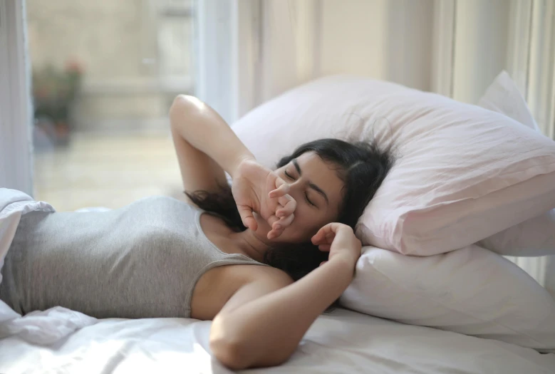 a woman laying on top of a bed next to pillows, pexels contest winner, happening, hand over mouth, profile image, white steam on the side, manuka