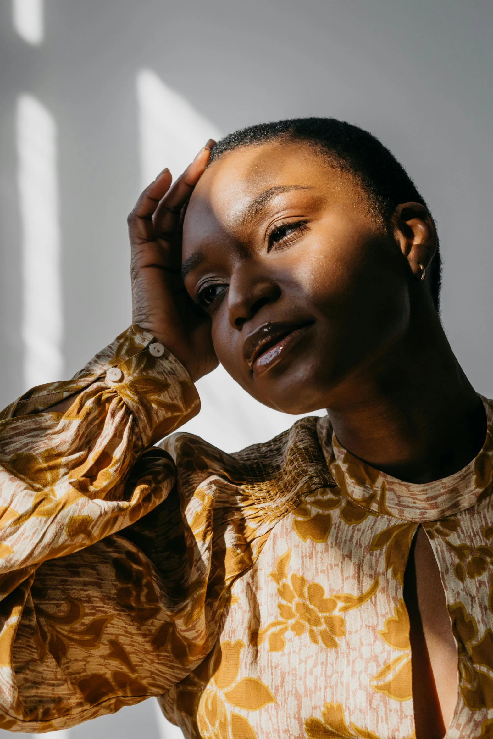 a woman standing in front of a window with her hands on her head, trending on pexels, wearing yellow floral blouse, dark skin tone, pondering, gold dappled light