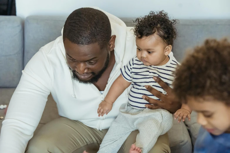 a man sitting on top of a couch holding a baby, by Arabella Rankin, pexels contest winner, varying ethnicities, sitting in a waiting room, caring fatherly wide forehead, jaylen brown