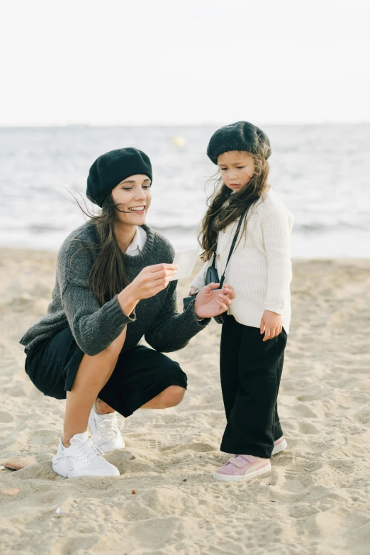 a woman and a little girl on the beach, a picture, by Lucia Peka, unsplash, wearing a french beret, black trousers, madison beer, black beanie