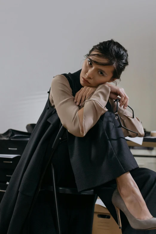 a woman sitting on top of a chair in a room, inspired by Tadashi Nakayama, trending on pexels, he is wearing a black trenchcoat, model is wearing techtical vest, wonbin lee, brown clothes