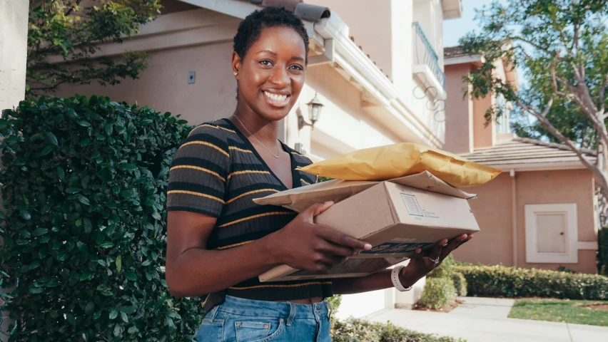 a woman holding a box in front of a house, dark skinned, ecommerce photograph, smiling, holding a gold bag