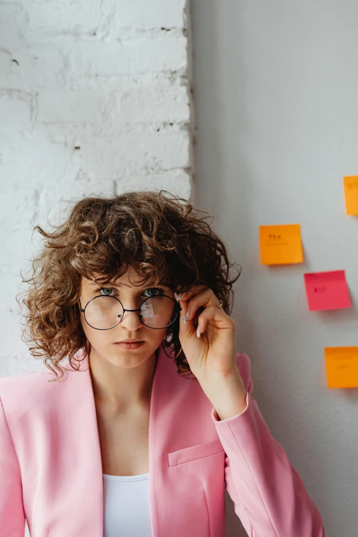 a woman sitting at a table in front of a laptop, trending on pexels, in square-rimmed glasses, curly haired, whiteboards, pink and yellow