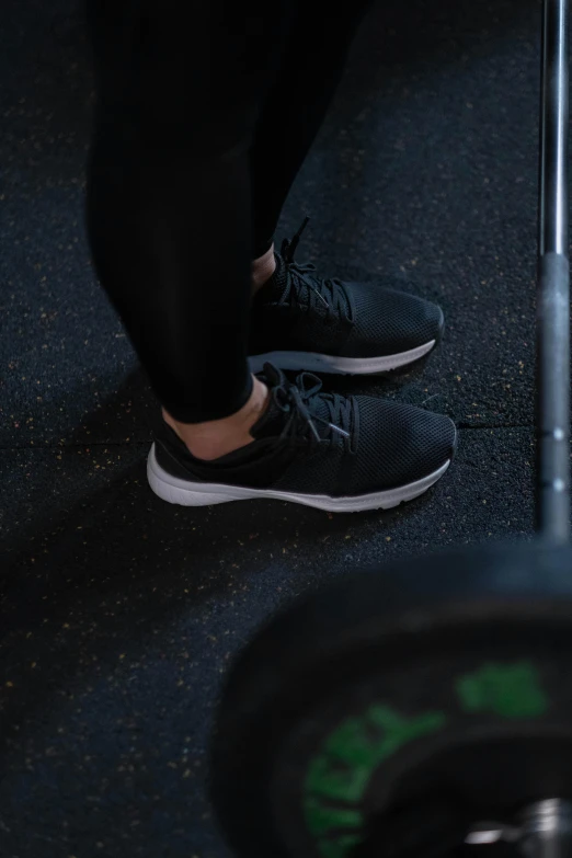 a person standing next to a barbell in a gym, running shoes, black on black, paved, background image