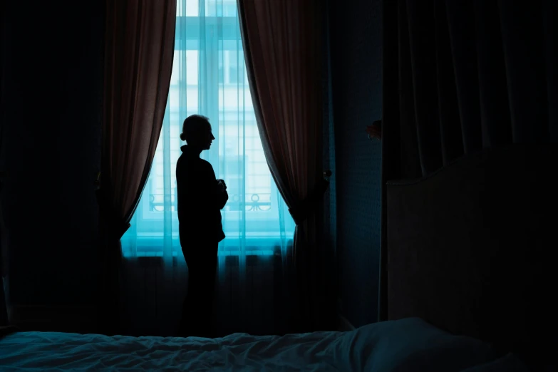 a person standing in front of a window in a dark room, a cartoon, inspired by Elsa Bleda, pexels contest winner, hotel room, blue backlight, black silhouette, depressing image