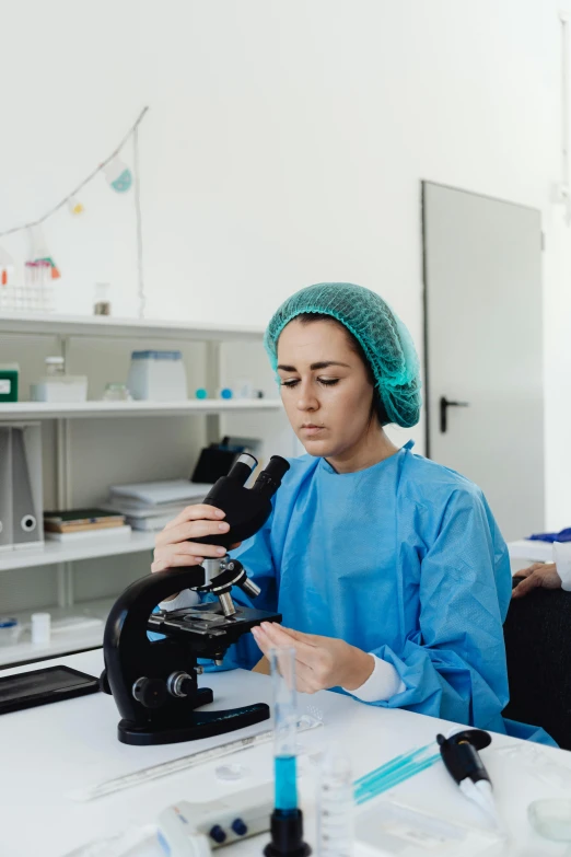 a woman sitting at a table looking through a microscope, a microscopic photo, trending on pexels, surgical gown and scrubs on, in a research facility, gif, paisley