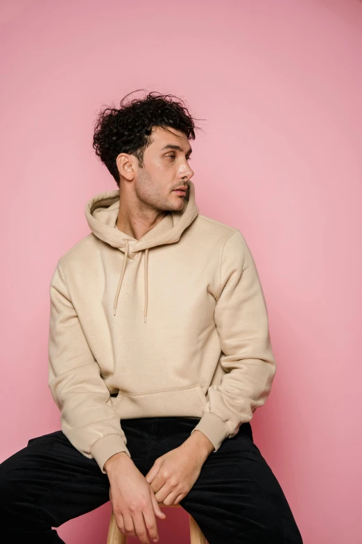 a man sitting on a stool in front of a pink wall, an album cover, inspired by Luca Zontini, trending on pexels, beige hoodie, headshot, riccardo scamarcio, blank background