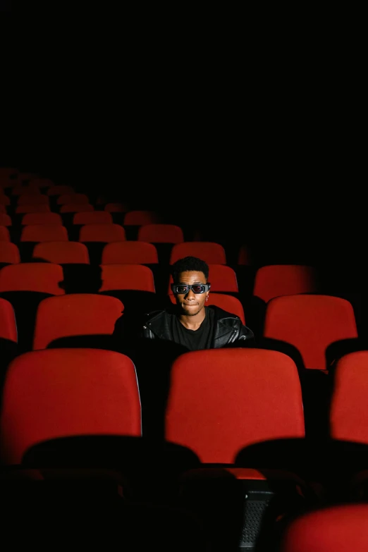 a man sitting in the middle of rows of red chairs, an album cover, pexels, wearing 3 d glasses, black teenage boy, movie poster with no text, [ theatrical ]