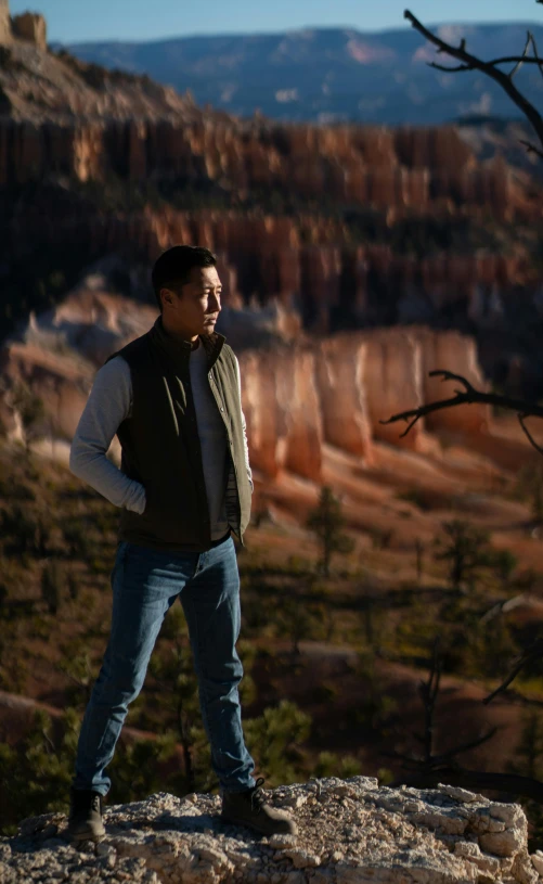 a man standing on top of a rock next to a forest, john cho, chiseled formations, behind the scenes photo, warm light