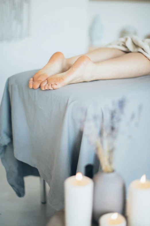 a woman laying on top of a bed next to candles, trending on pexels, massurrealism, close-up on legs, grey robes, sitting on a lab table, pastel'