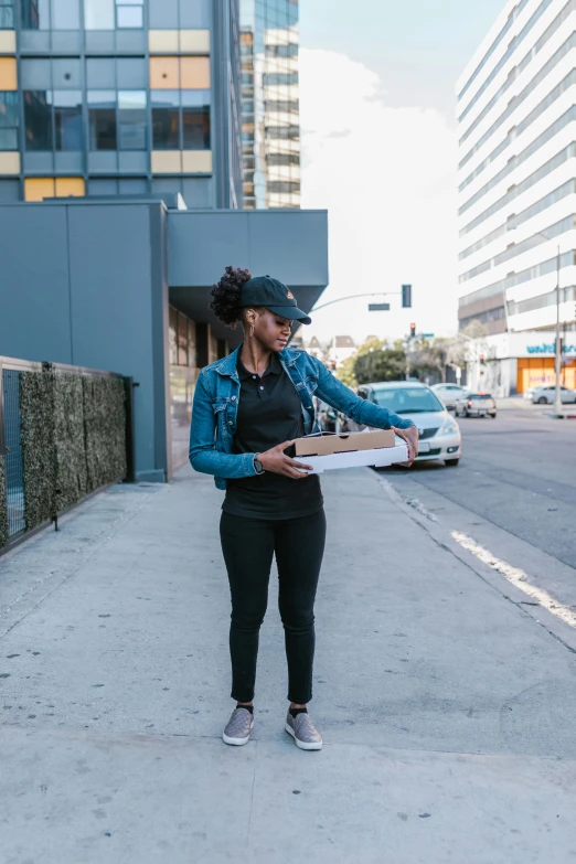 a woman standing on a sidewalk holding a box, sharing a pizza, wearing jeans and a black hoodie, hollywood standard, qiyana