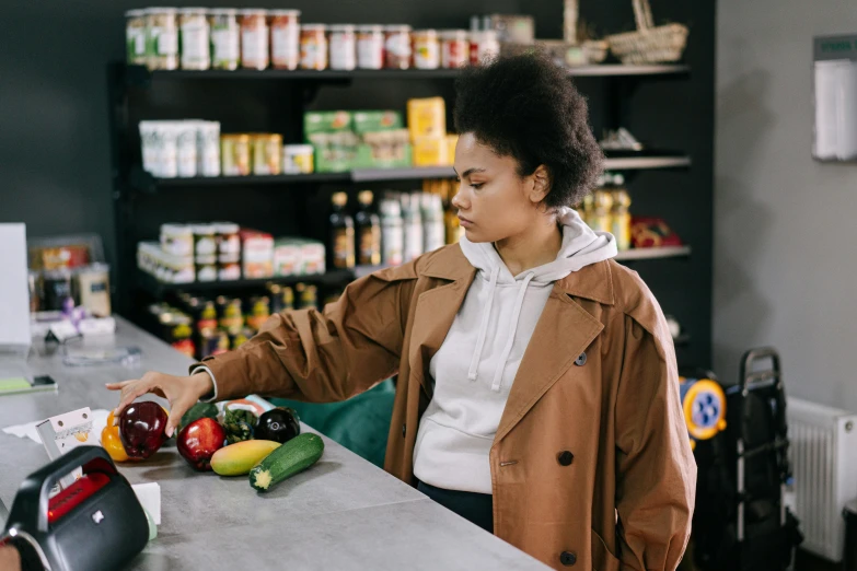 a woman standing at a counter in a grocery store, trending on pexels, tessa thompson, strange ingredients on the table, woman is in a trenchcoat, getting groceries