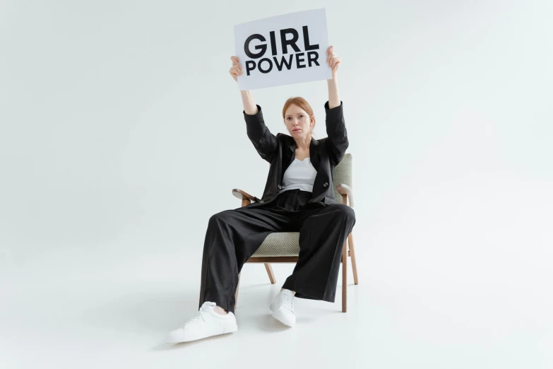 a woman sitting on a chair holding a sign, inspired by Harriet Powers, trending on pexels, feminist art, power girl, ellie bamber, super hero pose, black