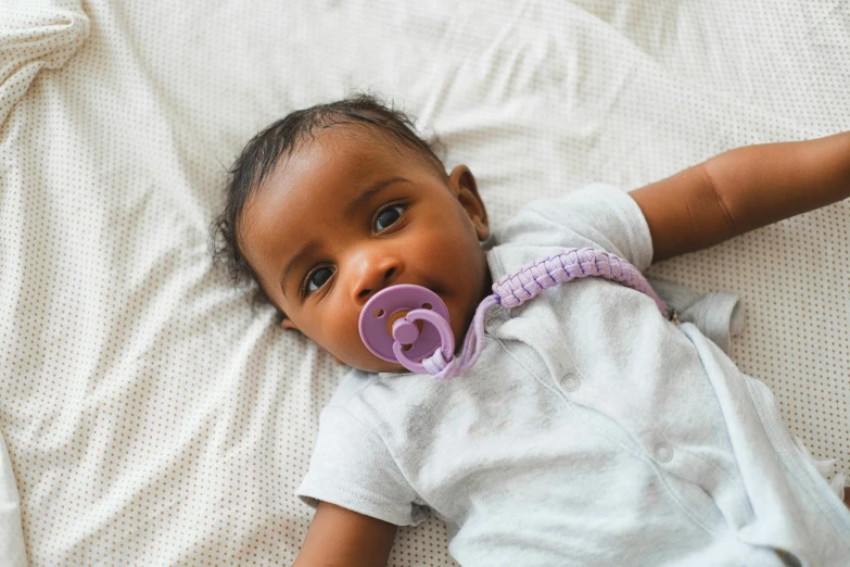 a baby laying on a bed with a pacifier in its mouth, by Nina Hamnett, pexels contest winner, happening, purple skin color, brown skinned, wearing collar on neck, on grey background