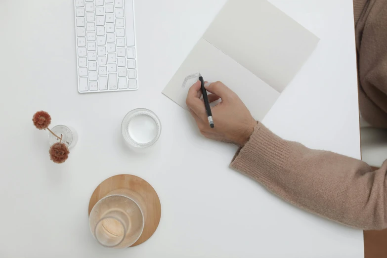 a person sitting at a desk writing on a piece of paper, pexels contest winner, white minimalistic background, background image, 9 9 designs, table in front with a cup