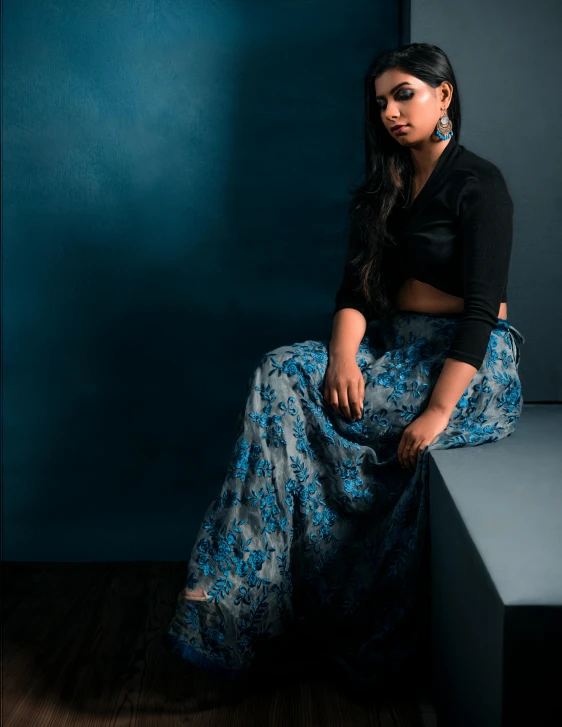 a woman sitting on a bench in front of a blue wall, an album cover, by Riza Abbasi, in a glowing skirt, in a dark studio room, formal wear, profile image