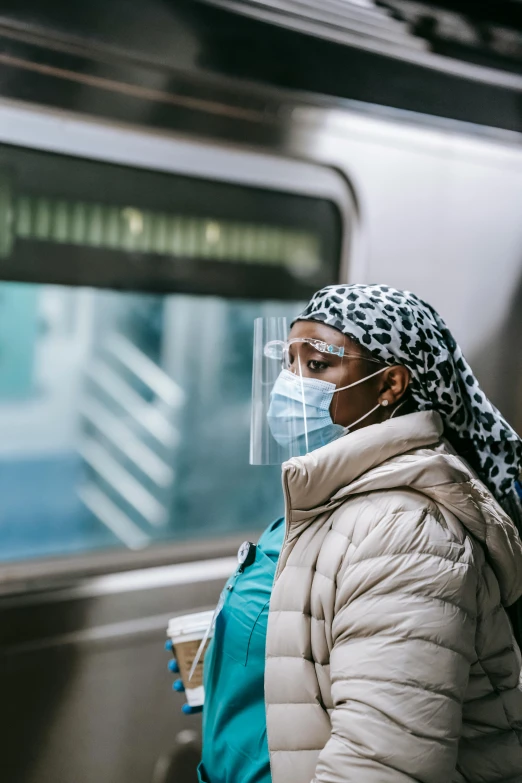 a woman wearing a face mask standing in front of a train, by Alison Geissler, pexels contest winner, humans of new york, slide show, aida muluneh, square