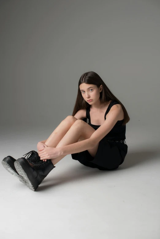 a woman sitting on the ground with her legs crossed, antipodeans, lorde, wearing tall combat boots, she is wearing a black tank top, concerned expression