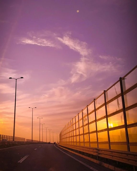 the sun is setting behind a fence on the side of the road, by Adam Rex, pexels contest winner, sky bridge, yellow and purple color scheme, jeddah city street, rainbow road